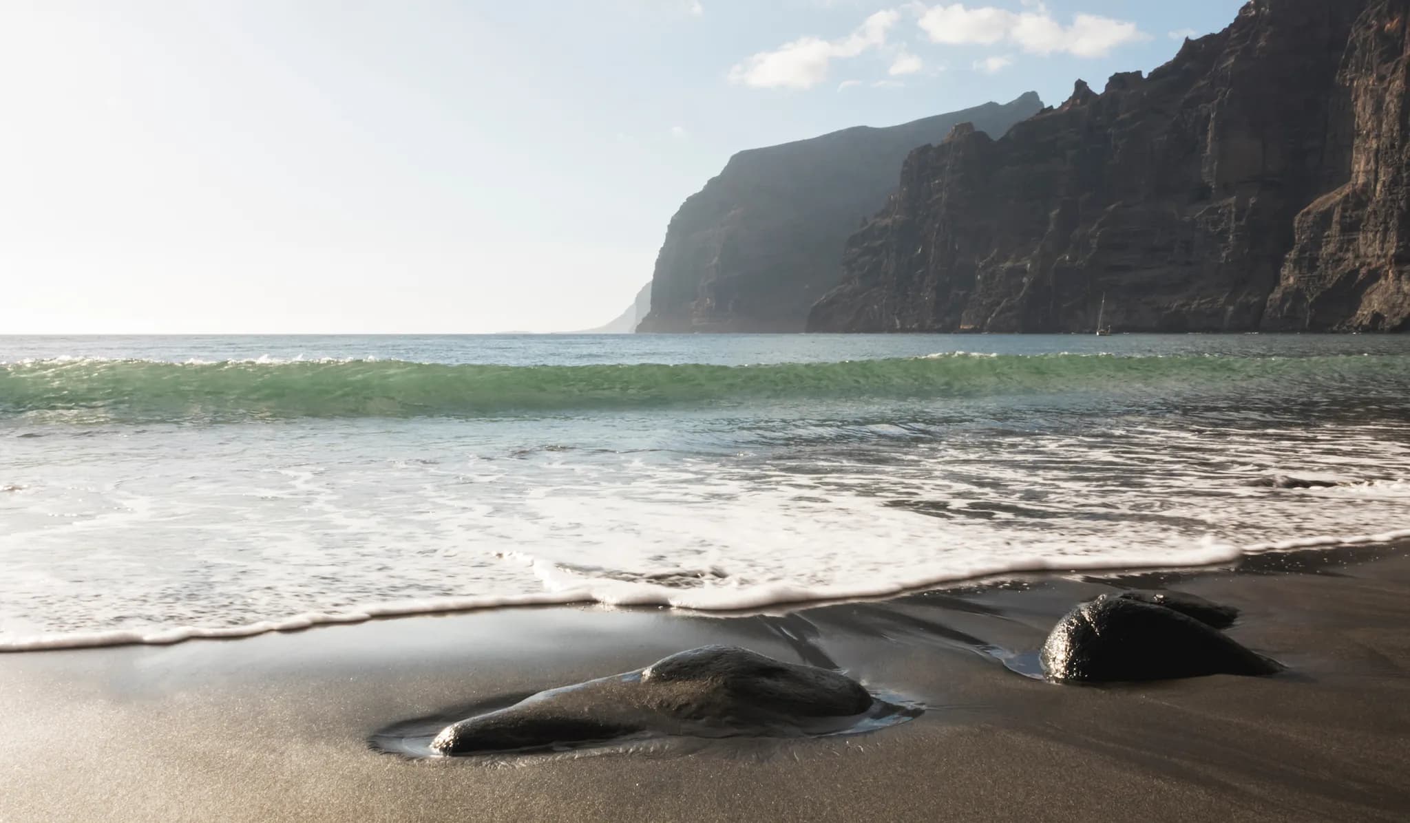 Vagues recouvrant le sable volcanique sombre et des galets noirs polis, avec des falaises en arrière-plan