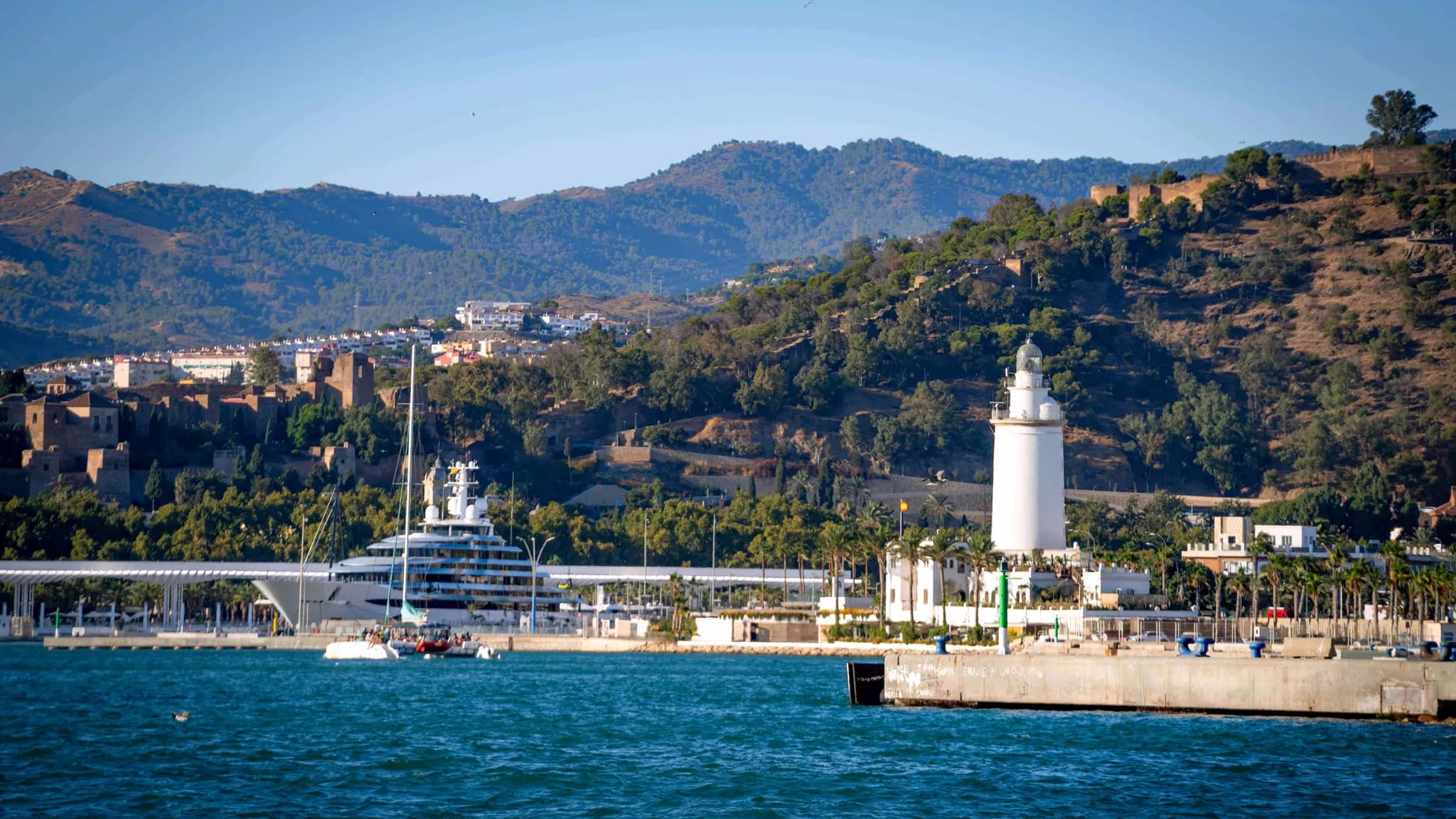 Le port de Malaga avec le phare de La Farola, un superyacht à quai et l'Alcazaba sur la colline boisée