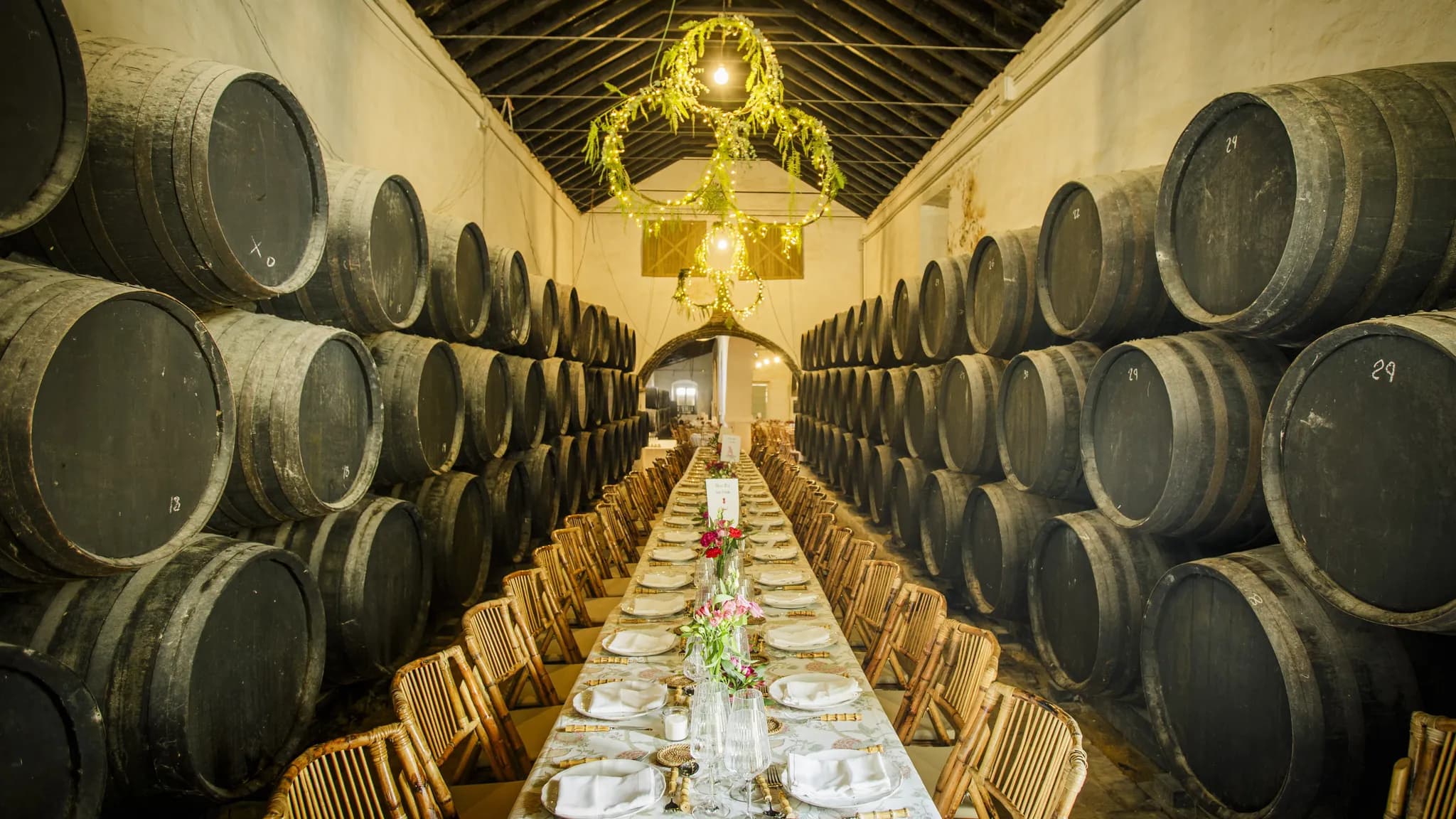 Une longue table de banquet dressée entre des rangées de fûts dans un chai, sous un lustre fleuri